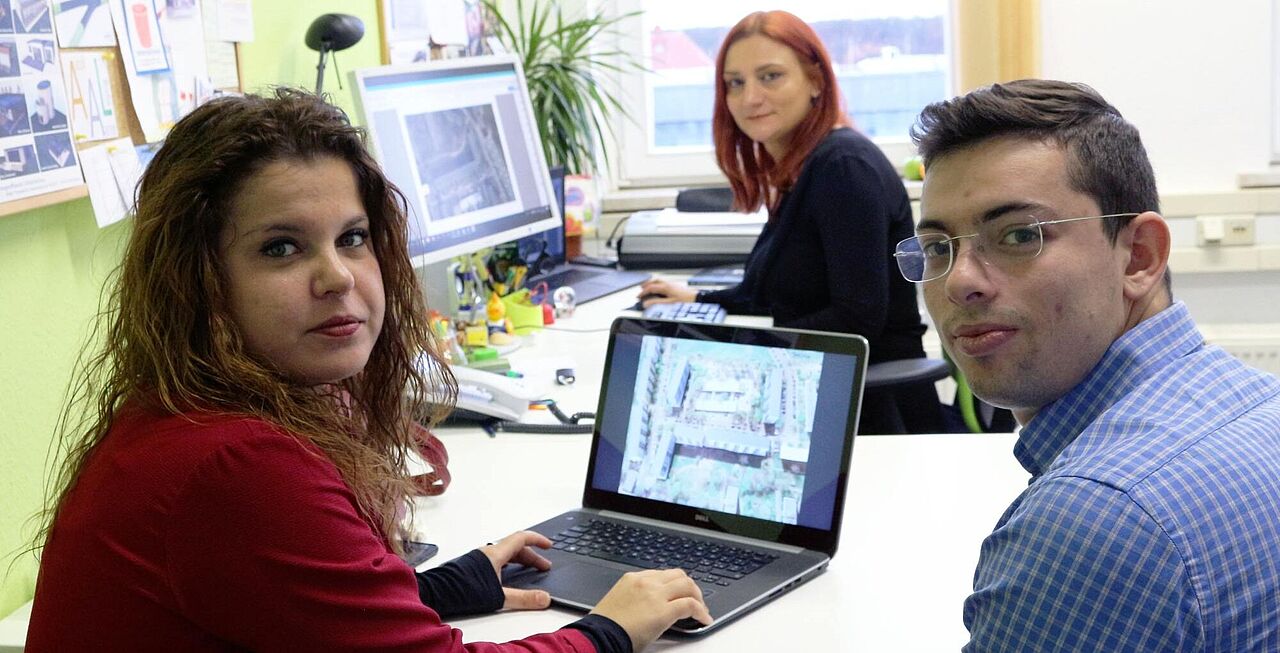 Aufnahme aus dem Büro der Professorin. Im Vordergrund sitzen die italienischen Doktoranden um einen Laptop herum, im Hintergrund sitzt Prof. Rossi an ihrem stationären Rechner. Beide Monitore zeigen Illustrationen einer Häuserzeile.
