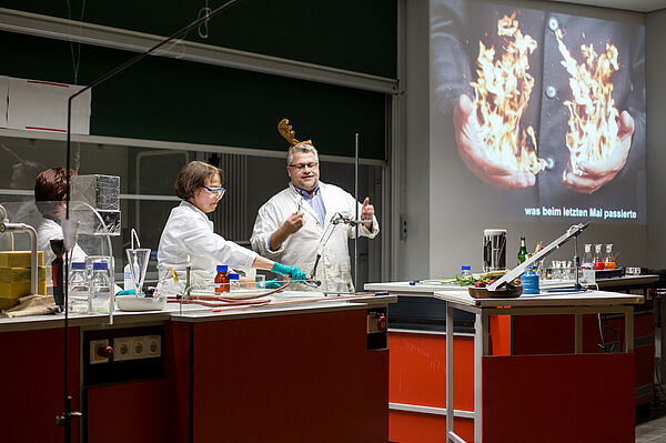 Versuchsanordnung der Weihnachtsvorlesung (Foto: R. Weinhold/HTWK Leipzig) Blick auf die Versuchsanordnung der Weihnachtsvorlesung. Auf dem Pult des Hörsaals stehen viele Chemielaborartikel, unter anderem ein Bunsenbrenner und zahlreiche Fläschchen. Uta Greif erwärmt mit einen weißen Feststoff in einem Reagenzglas, Lutz Engisch steht dahinter und schaut zu.
