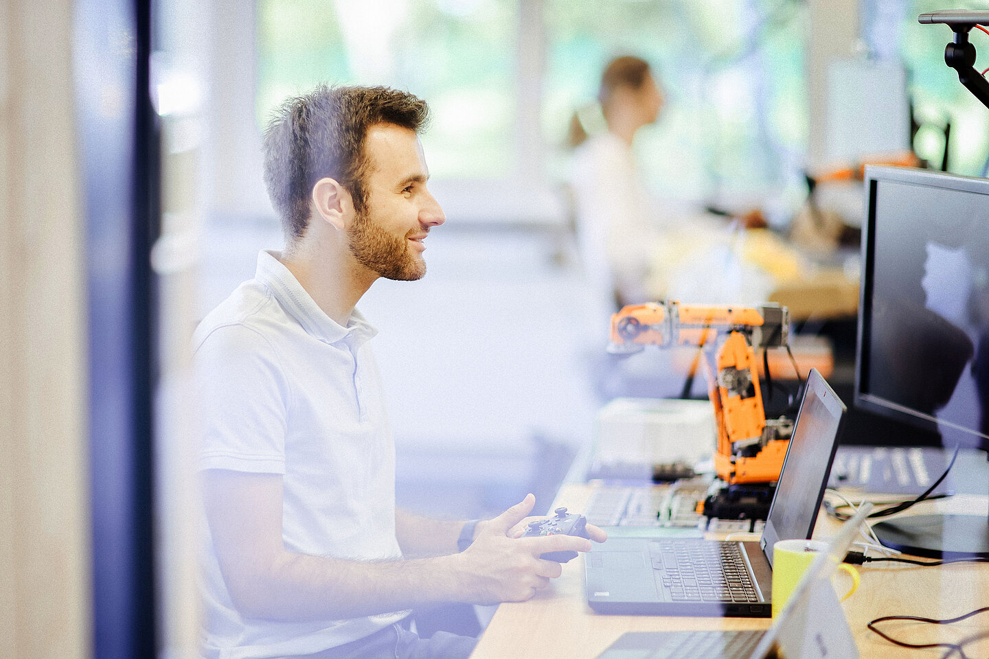 Mann mit Gamecontroller steuert einen kleinen orangefarbenen Roboterarm am Schreibtisch mit Laptop und Monitor in einem modernen Büro.