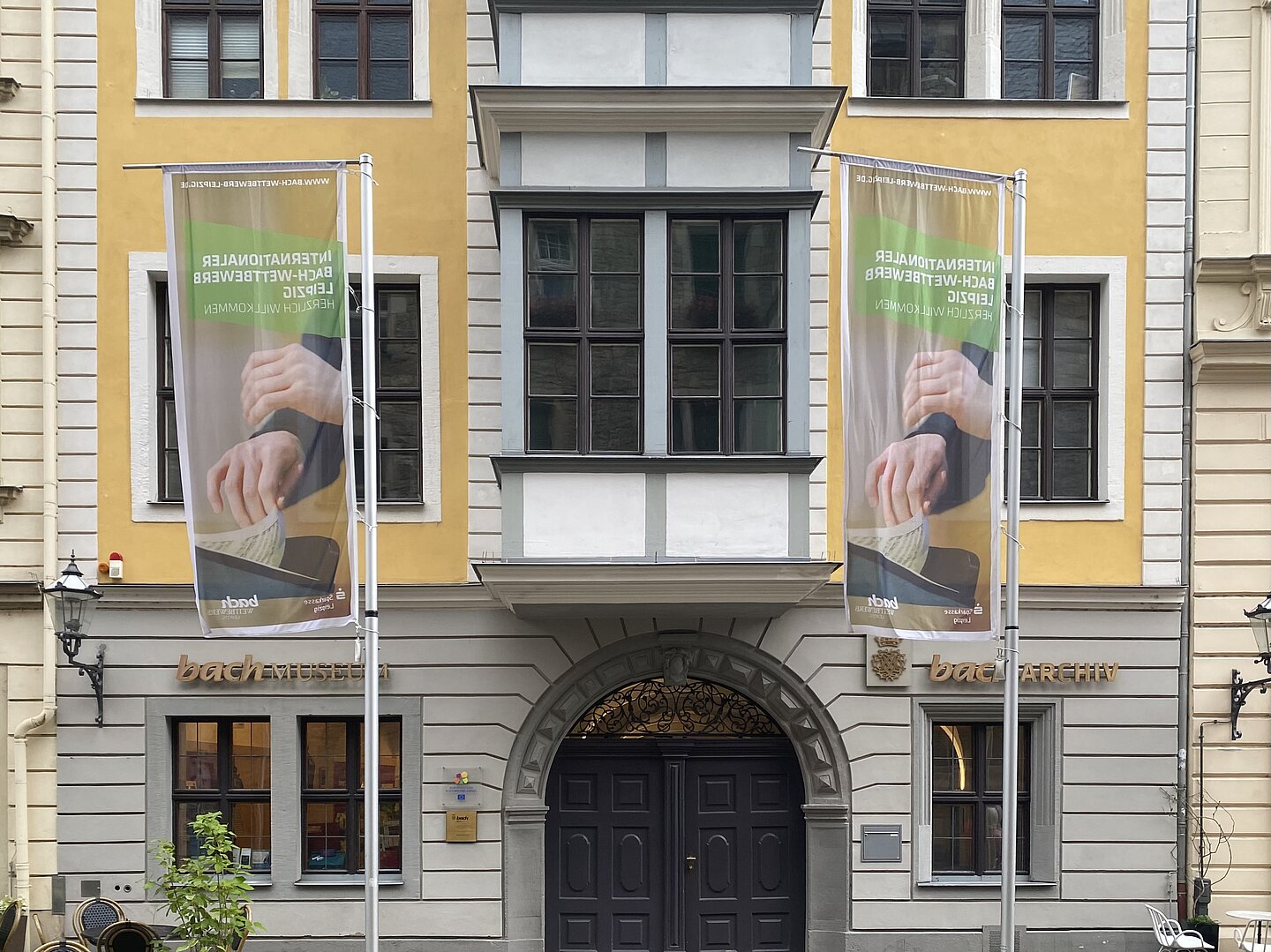 Bach-Museum und Bach-Archiv Leipzig Das Bild zeigt die Fassade des Bach-Museums und Bach-Archivs. Das Gebäude hat große Fenstern und einen auffälligen Eingangsbereich mit einem dunklen Holztor. Zwei Fahnenmasten stehen vor dem Gebäude, an denen Banner hängen. Die Fassade ist in gelben und grauen Farbtönen gehalten. Der Schriftzug "bachMUSEUM" und "bachARCHIV" ist an der Vorderseite des Gebäudes zu sehen.