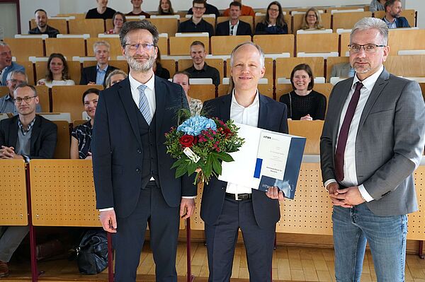 Gruppe von drei Herren mit Urkunde und Blumen vor Publikum in Hörsaal