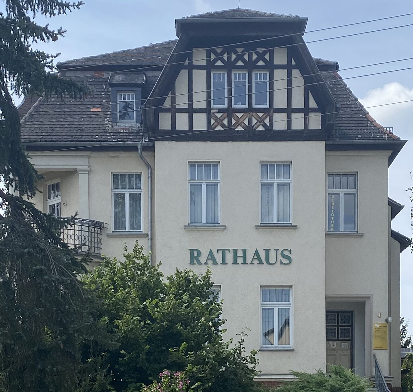 Sie Hochgeladenes Bild beschreibe Copilot Das Bild zeigt ein Gebäude mit der Aufschrift "RATHAUS" auf der Fassade, was auf ein Rathaus hinweist. Das Gebäude kombiniert traditionelle und moderne Elemente: Es hat ein Satteldach, mehrere Fenster und einen kleinen Balkon auf der linken Seite. Rechts am Gebäude befindet sich ein Schild mit der Aufschrift "BIBLIOTHEK", was auf eine Bibliothek hinweist. Um das Gebäude herum gibt es Grünflächen.
