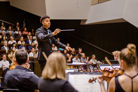 Während der feierlichen Immatrikulation spielt das HTWK Orchester im Gewandhaus. Man sieht meherer Menschen von hinten ihr Intrument spielen, vor ihnen steht der Dirigent.