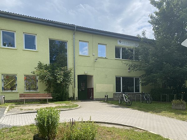Phyllodrom e. V. Ein zweistöckiges Gebäude mit grüner Fassade. Davor gibt es einen gepflasterten Weg mit kleinen Grünflächen daneben. Vor einem Fenster des Gebäudes steht eine Bank.