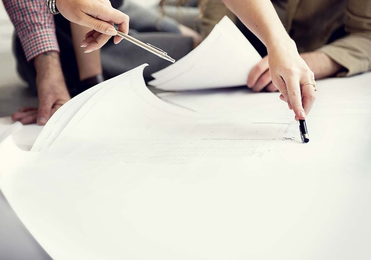 Auf dem Tisch liegt ein Plan, welcher neunzig Prozent des Bildes ausmacht. Hände zeigen mit Stiften auf den Plan.