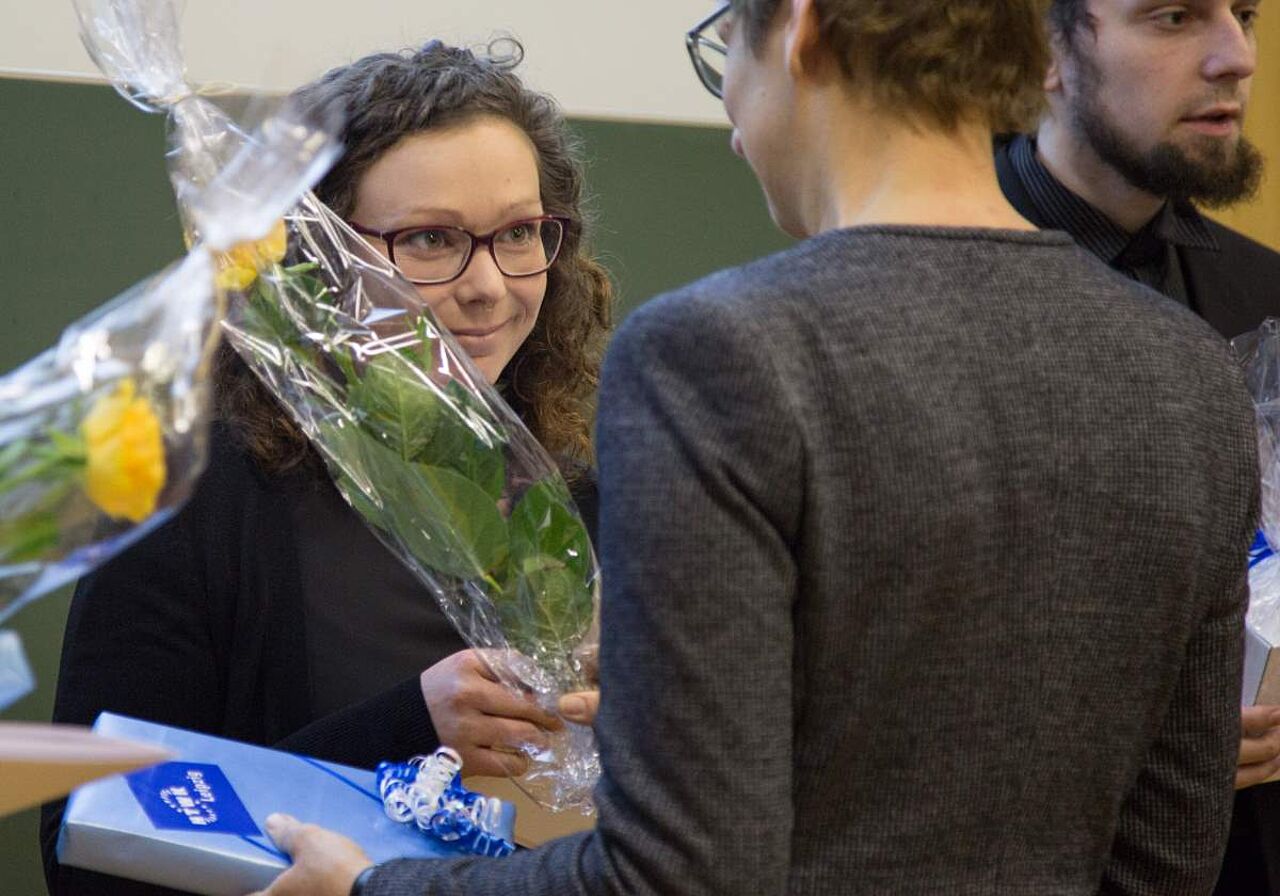Ein Person reicht einer Frau ein Blumenstrauß und ein Geschenk. Die Frau hat eine Brille und lockige braune Haare. Von der Person ist nur der Rücken und die Hände zu sehen. Daneben erknennt man das Gesicht eines Mannes.