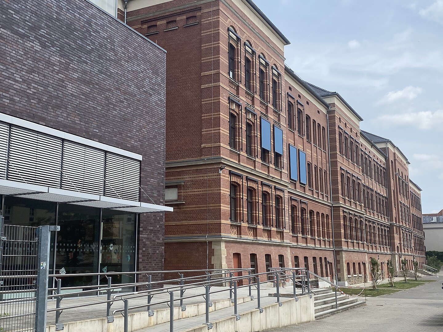 Werner-Heisenberg-Schule Das Bild zeigt ein großes, mehrstöckiges Backsteingebäude. Die Fassade besteht aus rotem Ziegelstein und ist durch regelmäßige Fensterreihen strukturiert. Im Vordergrund ist ein moderner Anbau mit dunkler Backsteinverkleidung zu sehen, der sich durch große Fensterfronten auszeichnet. Eine Rampe führt zum Eingangsbereich des Anbaus. Die Architektur verbindet klassische und moderne Stilelemente.