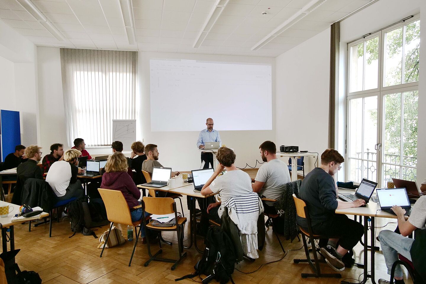 (Foto: Suse Eckhart) HTWK-Professor Gerold Bausch steht vorn, via Beamer wird ein Bild an die Wand projiziert. Die Studierenden sitzen an Tischen und hören zu.
