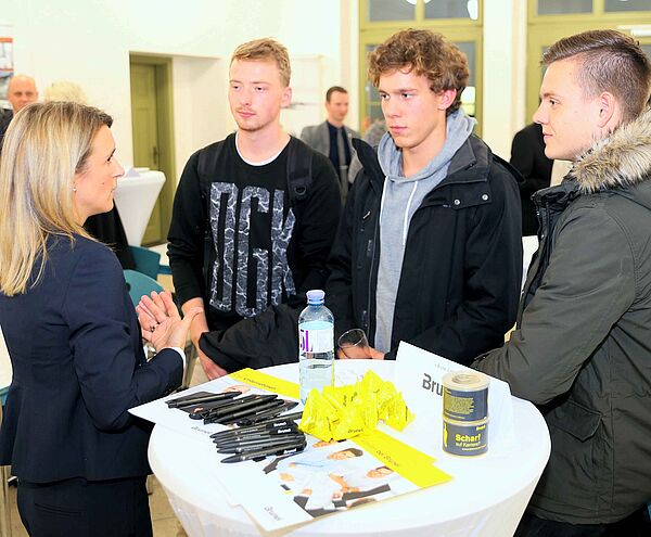 Persönlicher Kontakt zwischen Unternehmen und Studierenden der HTWK Leipzig bei "Unternehmen zu Gast". Eine Frau erklärt drei Studierenden an einem Stand ihr Unternehmen. Sie stehen um einen Tisch herum auf den Flyer liegen.