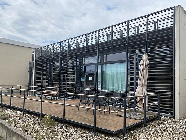 Klinikum St.Georg Lesecafe Terasse Das Bild zeigt die Außenseite eines Gebäudeabschnitts mit Terasse. Die Fassade des Hauses setzt sich aus großen Glasfenstern zusammen. Vor diesen erstreckt sich ein schwarzes gitterähnliches Gerüst, welches über die Dachhöhe hinausragt und eine Dachterasse bildet. Die waagerechten Stangen des Gerüstes, haben einen geringen Abstand zueinander und bieten somit zusätzlichen Sichtschutz. An zwei Glasscheiben setzten sie jedoch aus, um einen Durchgang und Ausblick zu schaffen. Auf der zentralen Scheibe steht oben in weißen Großbuchstaben "Lesecafe" geschrieben. Darunter bedindet sich die ebenfalls gläserne Tür. Von innen ist an dem danebenliegenden Fenster ein weißes Rollo angebracht, dass die Sicht nach drinnen versprerrt. Die Terasse vor dem Gebäudewürfel grenzt sich mit einem Holzlattenboden von dem darunterliegenden Kiesboden ab. Während auf der linken Seite lediglich eine Holzbank steht, befinden sich rechts fünf, in einem Kreis aufgestellte, schwarze Stühle. Ganz am Rand daneben wurde ein weißer Sonnenschirm aufgestellt.