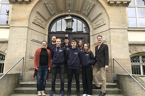 Studentisches Projektteam, sowie Projektmitarbeiter:innen und Professor stehen gemeinsam auf der Treppe vor dem Gutenbergbau.