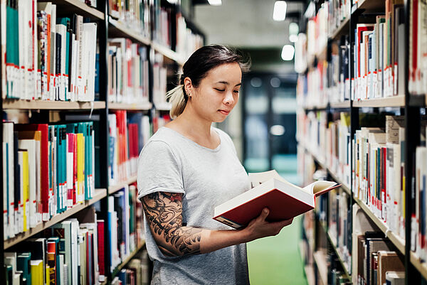 Eine Studentin blättert zwischen Bibliotheksregalen in einem Buch.