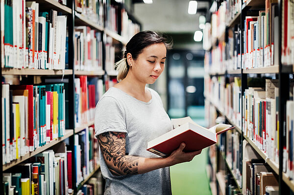Eine Studentin blättert zwischen Bibliotheksregalen in einem Buch.