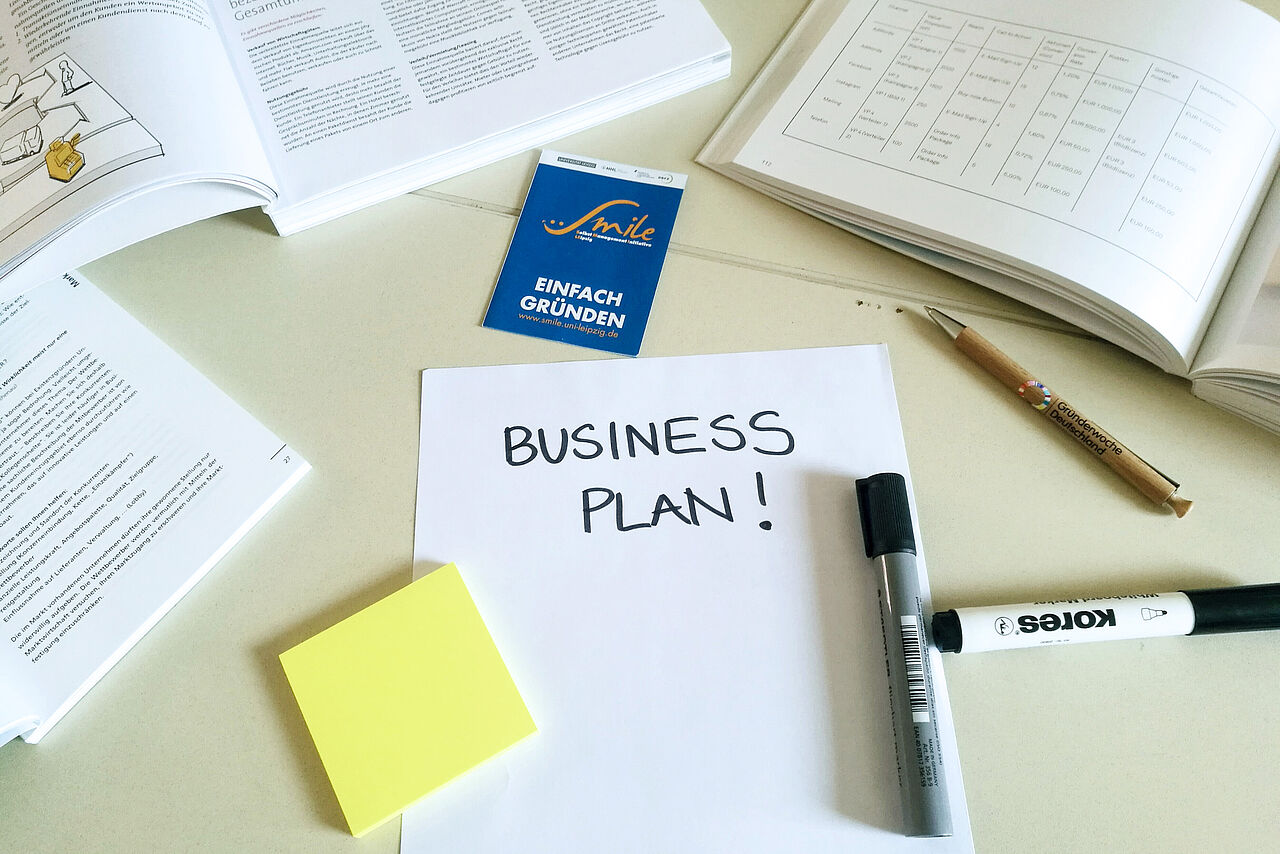 Auf einem Schreibtisch liegen verschiedene Notizbücher und Stifte. Auf einem weißen Blatt Papier ist "Buisiness Plan" geschrieben