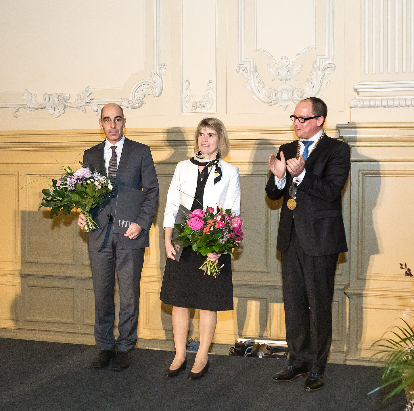 Prof. Mark Mietzner (rechts) mit Prof. Ralf Thiele, Prorektor Forschung, und Prof. Barbara Mikus, Prorektorin Bildung. (Foto: Swen Reichhold/HTWK Leipzig) Drei Personen mit Blumen auf Bühne: zwei Männer und in der Mitte eine Frau.