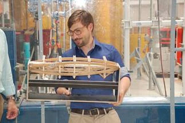 Eine Person hält ein Modell aus Holz in der Hand und blickt darauf. Im Hintergrund ist ein Labor
