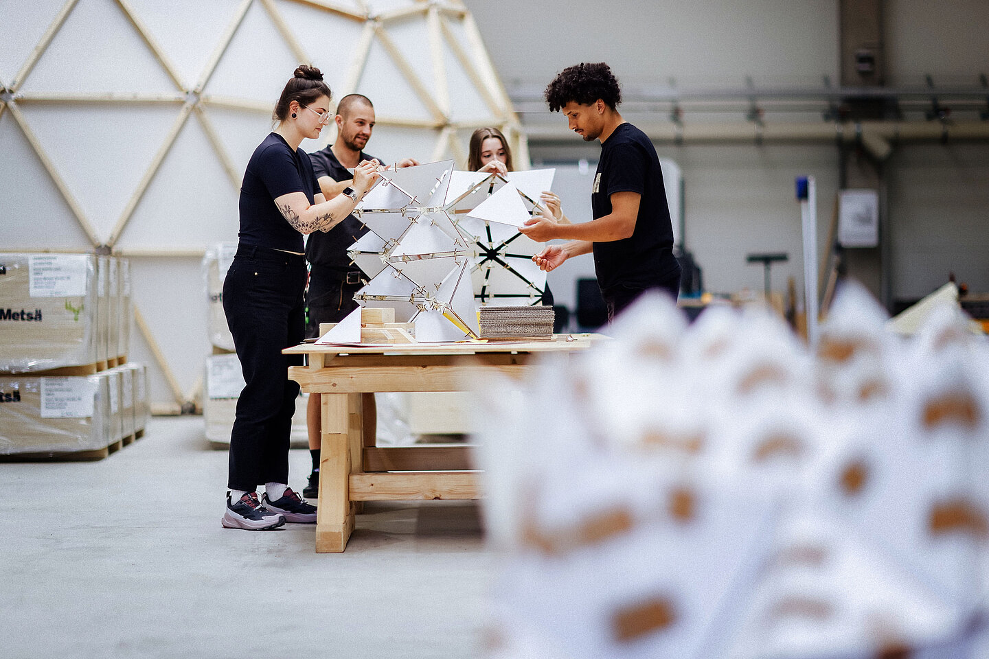 Vier Personen arbeiten an einer geometrischen Struktur aus Dreiecken auf einem Holztisch in heller Werkhalle.