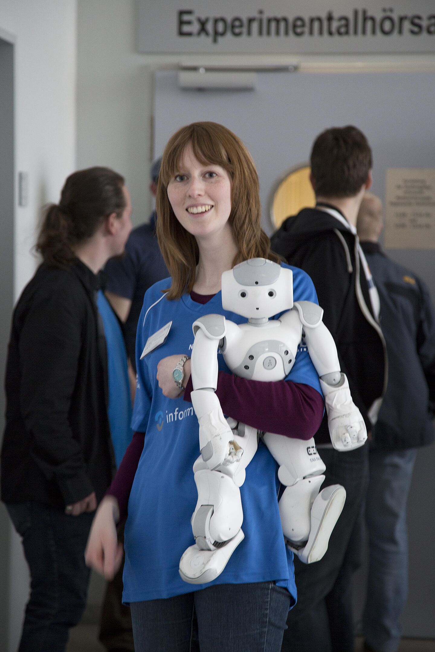 Anne Wissing mit Nao-Roboter „Megamind“ (Foto: Andreas Schröder/HTWK Leipzig) Anne Wissing trägt ein blaues Trikot der HTWK Leipzig und hält den Roboter "Megamind" im Arm. Hinter ihr sind ein paar Menschen von hinten zu sehen. Sie lächelt in die Kamera.