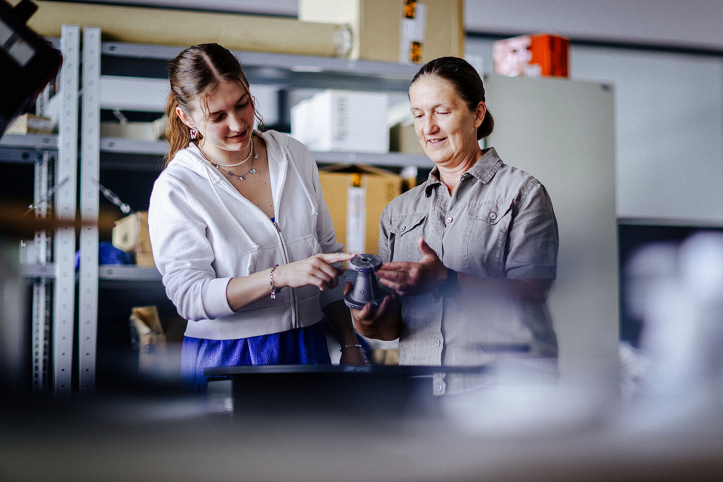 Zwei Frauen stehen vor Regalwand mit Kartons; eine hält ein graues Bauteil in den Händen und erklärt der anderen etwas dazu.