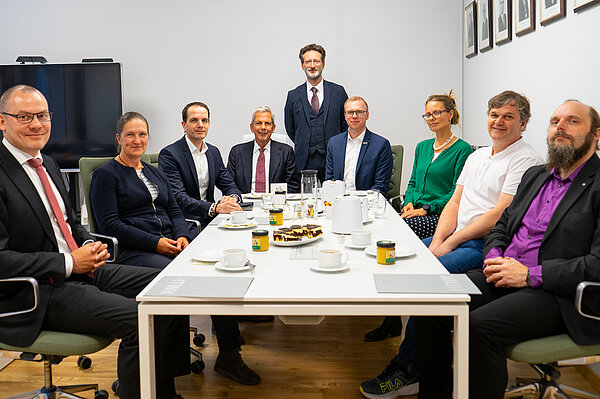 mehrere Personen sitzen als Gruppe an einem Tisch und lächeln in die Kamera