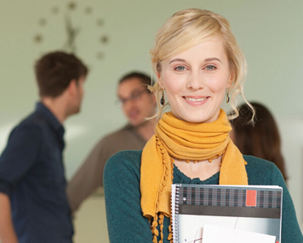 Eine junge Frau ist vorne im Bild. Sie lächelt in die Kamera und hat einen Schreibblock an ihre Brust gedrückt. Im Hintergrund sieht man drei weitere Studierende miteinander reden.