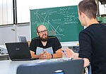 Ein Student mit Glatze und Bart sitzt an einem Tisch mit einem Laptop vor sich. Er schaut zu einem andern Studenten und erklärt diesem etwas. Im Hintergrund ist eine Tafel.