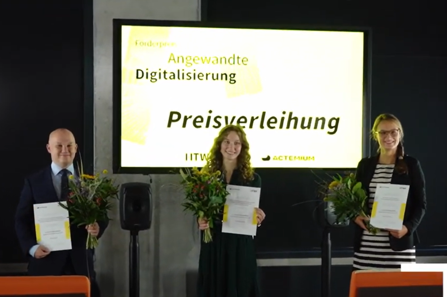 Ein Mann und zwei Frauen mit Urkunden in der Hand stehen vor einer Leinwand