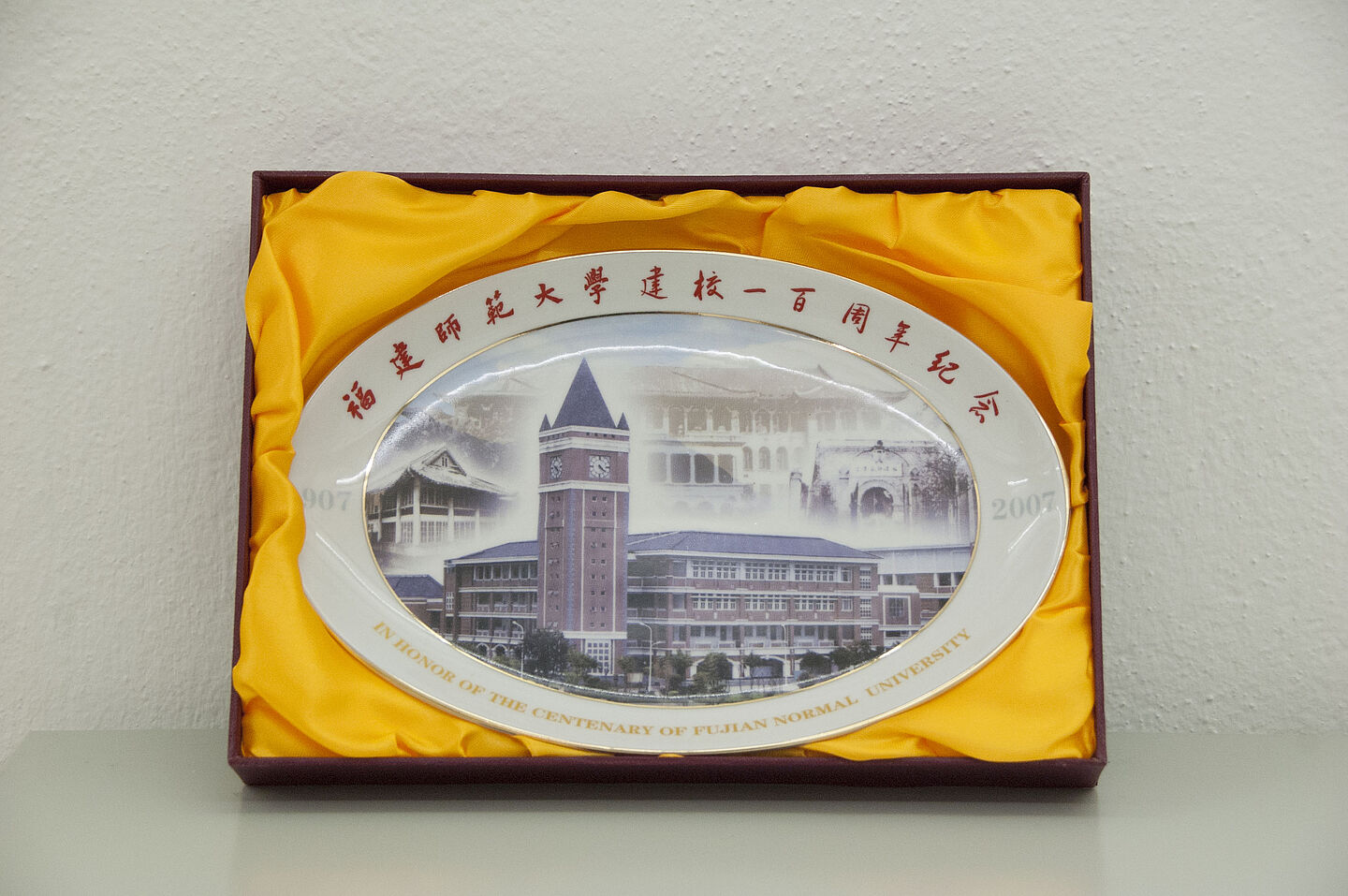 Chinesischer Teller aus dem HTWK-Archiv (Foto: HTWK Leipzig) Zu sehen ist ein chinesischer, ovaler Teller aus weißem Porzellan, der in einer ausgefütterten Schatulle liegt. Auf dem Teller ist das Bild eines alten Universitätsgebäudes mit Uhrturm zu sehen. Unter dem Bild steht: "In Honor of the Centenary of Fujian Normal University". Über dem Bild stehen chinesische Schriftzeichen.