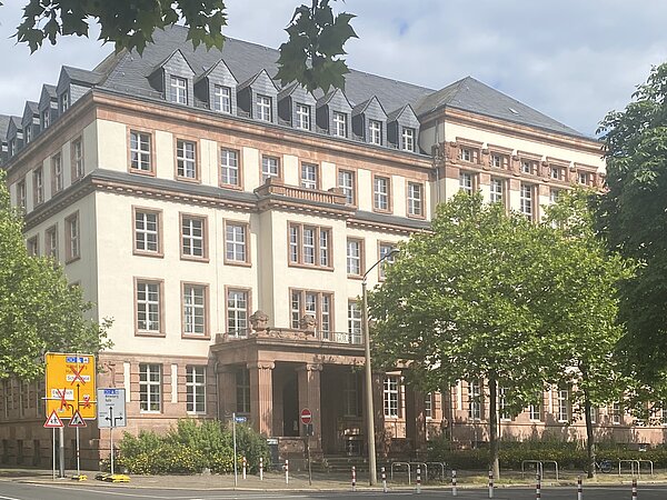 Ein fünfstöckiges Altbaugebäude von Außen mit beiger Fassade und grauem Dach. Der Eingangsbereich ist überdacht. Vor dem Gebäude stehen Bäume.