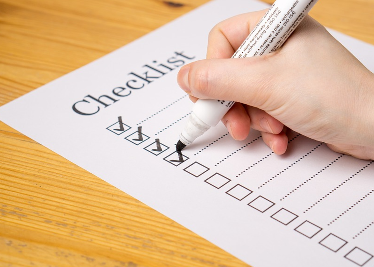 Es ist eine Hand zu sehen, in der ein Stift gehalten wird. Die Person hakt Punkte auf einer Checkliste ab.