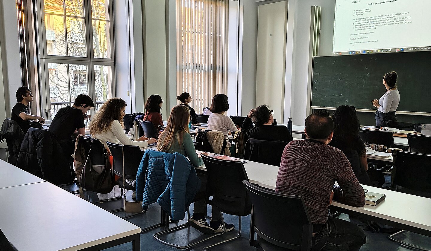 Das Bild zeigt einen Ausschnitt aus einer Unterrichtsstunde. Kursteilnehmenden sitzen an der Schulbank und die Dozentin zeigt auf eine virtuelle Tafel und erklärt etwas.