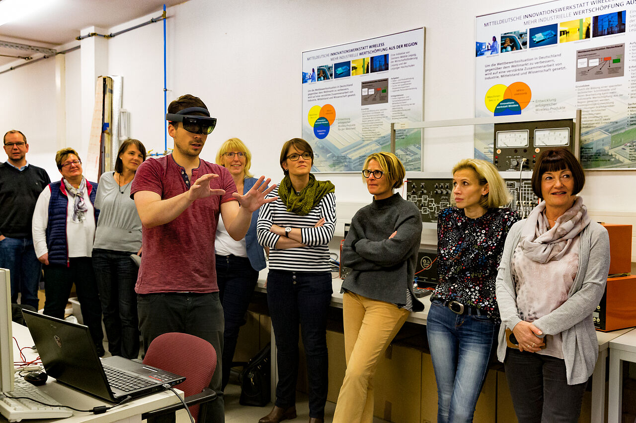 Teilehmer einer Fortbildung stehen um einen Studenten herum, der eine VR-Brille trägt und in die Luft tippt.