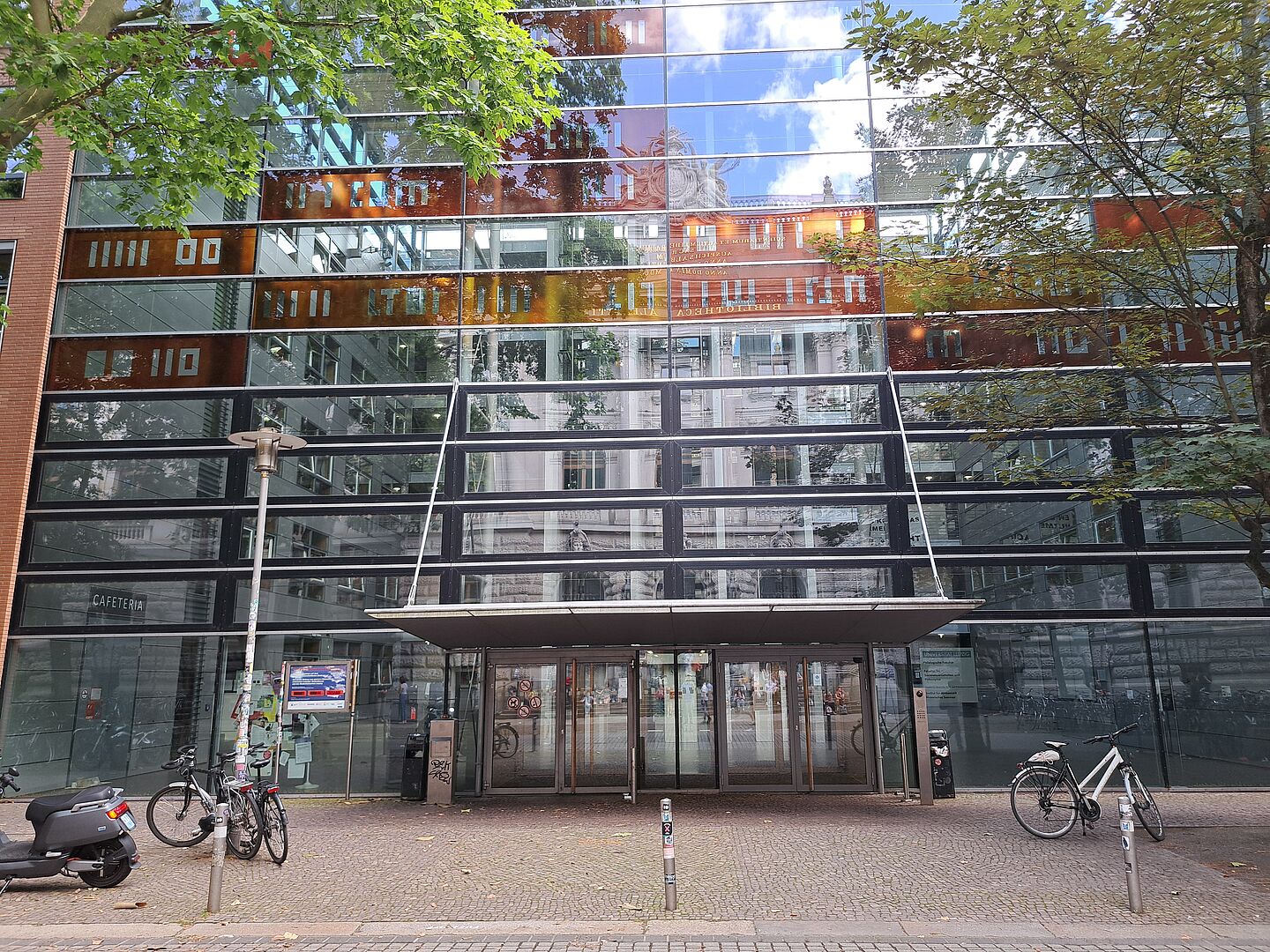 Das Bild zeigt das Québec-Studienzentrum an der Universität Leipzig, ein modernes Gebäude mit einer markanten Glasfassade, die das umliegende städtische Leben und die Baumkronen widerspiegelt. Das Design ist zeitgenössisch, mit klaren Linien und einer strukturierten Anordnung von Fenstern und Paneelen. Über dem Eingang ist ein Vordach angebracht, und die Türen sind ebenfalls aus Glas. Vor dem Gebäude befinden sich Fahrradständer, ein Motorroller.