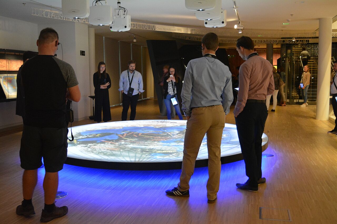 Teilnehmer einer Besuchergruppe schauen auf ein beleuchtetes kreisrundes Modell am Boden.