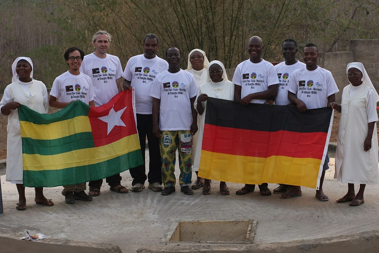 Gruppenfoto mit Fahnen beider Nationen (Togo und Deutschland). Zu sehen sind knapp ein Dutzend Menschen nebeneinander, darunter deutsche Planer, einheimische Arbeiter und Ordensschwestern.