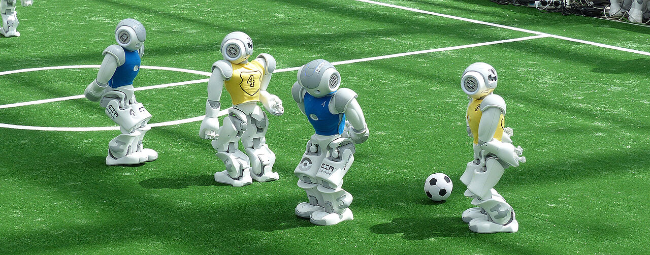 Vier Roboter, jeweils zwei von jedem Team, spielen auf einem Kunstrasen Fussball.