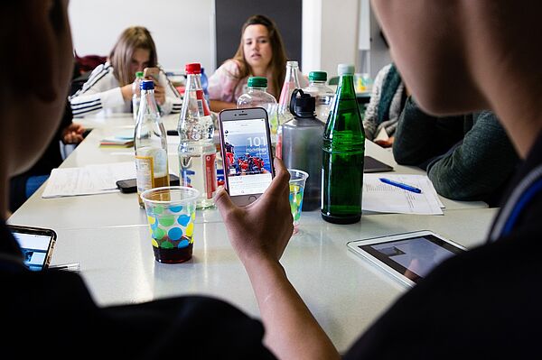 ein Tisch mit mehreren Person, zwei halten ein Smartphone und schauen rein