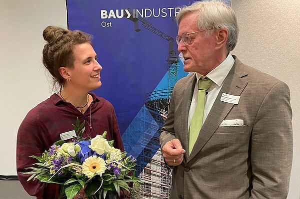 links steht eine Frau und rechts ein Mann, sie schauen sich an. Die Frau hält einen Blumenstrauß.