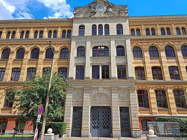 Ein großes, alt verziertes Gebäude mit teils beiger und teils gelber Fassade mit roten Akzenten. Der Haupteingang besteht aus einer große, blauen Tür mit zwei kleineren Eingängen an den Seiten.