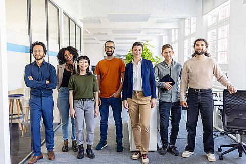 Eine multikulturelle Gruppe von jungen Erwachsenen steht in einem Büro und alle schauen frontal in die Kamera.