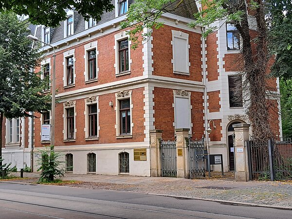 Mehrstöckiges rotes Backsteingebäude mit weißen Verzierungen. Der Eingang befindet sich hinter einem geöffneten Tor.