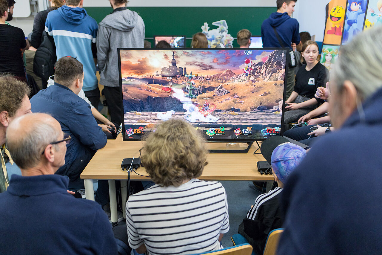 Mehrere Menschen sitzen um einen Monitor herum, auf dem ein Computerspiel läuft