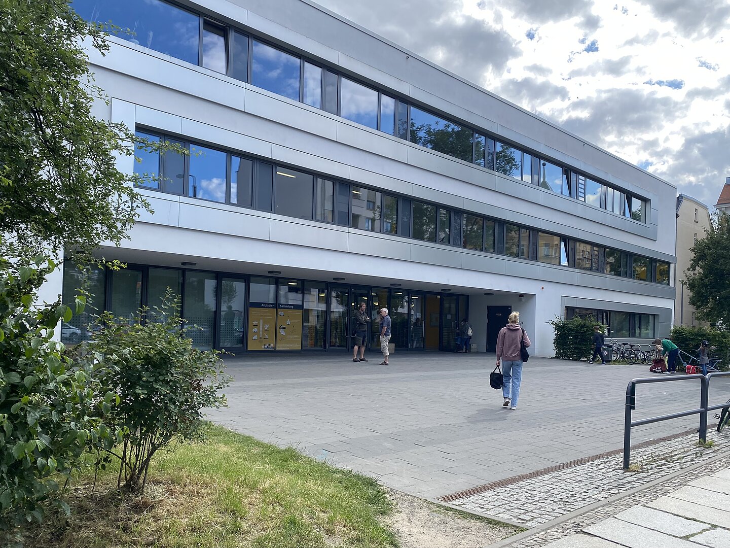 Kurt-Masur-Schule Das Bild zeigt ein modernes, mehrstöckiges Gebäude mit großen Fenstern und einem flachen Dach. Es hat drei sichtbare Stockwerke und einen Haupteingang im Erdgeschoss. Der Bereich vor dem Gebäude ist gepflastert und links sind einige Grünflächen und Büsche zu sehen. Außerdem sind Fahrräder in der Nähe des Eingangs auf der rechten Seite geparkt.