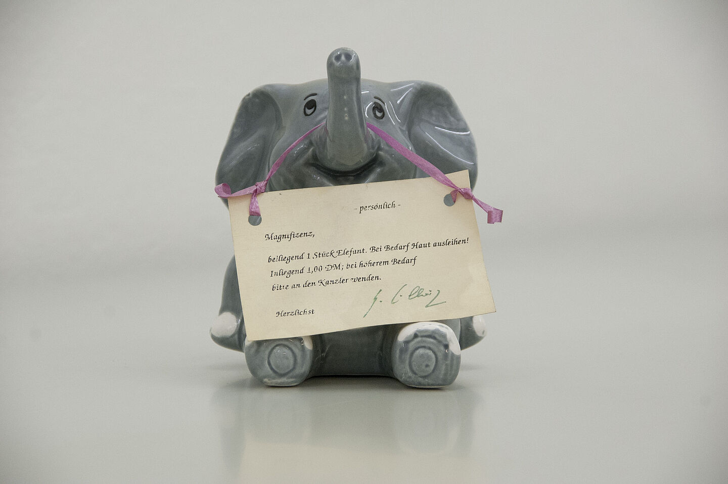Elefantenspardose aus dem HTWK-Archiv (Foto: HTWK Leipzig) Zu sehen ist ein kleiner Elefant aus grauem Porzellan, der auf dem Boden sitzt. An seinem ausgestreckten rüssel hängt ein Papierschild mit einer persönlichen Widmung und Unterschrift.
