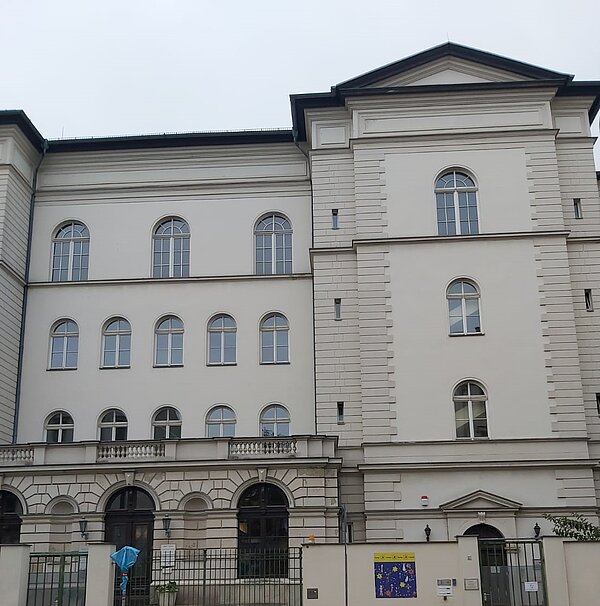 Ein hellbeiges, alt verziertes Gebäude mit Rundbogenfenstern. Vor dem Gebäude ist eine Mauer mit Metalltoren.