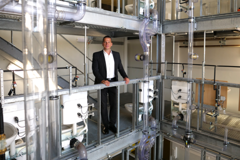 Erweiterung des Abwasserturms Nieperbau Mann in Anzug steht auf einer Metallplattform in einem Sanitärlabor. Um ihn herum sind durchsichtige Rohre, Toiletten und Waschbecken in mehreren Etagen