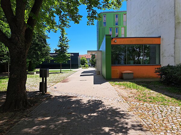 Ein Gebäude mit schmutzig weißer Fassade und einem kleinen orange-grünen Vorbau, in dem sich der Eingang befindet. Auf der Fassade sieht man den Teil eines Schriftzugs. Das Gebäude steht auf der rechten Seite eines Weges, weitere Gebäudekomplexe befinden sich zu beiden Seiten dieses Weges.