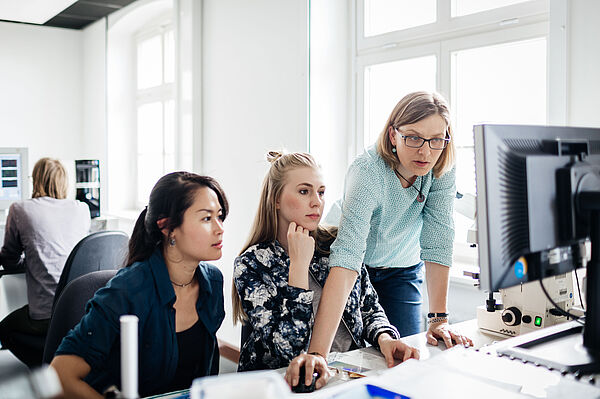 Dozentin zeigt zwei Studentinnen am Schreibtisch etwas auf dem Monitor