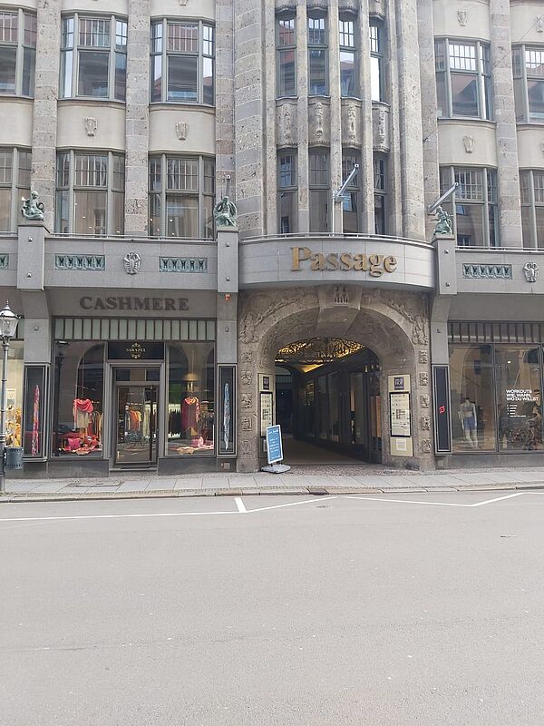 Das Bild zeigt den leicht gewölbten Eingang eines Einkaufs- und Geschäftspassage-Gebäudes in einem städtischen Umfeld. Über dem Eingang prangt das Wort „Passage“ in goldener Schrift. Links und rechts neben dem Eingang befinden sich Schaufenster von Geschäften. Die Architektur des Gebäudes ist markant und scheint von einem klassischen Stil inspiriert zu sein, mit dekorativen Elementen aus Stein, symmetrischen Fenstern und kleinen Verzierungen, wie zwei sitzenden Figuren auf beiden Seiten oberhalb des Eingangs.