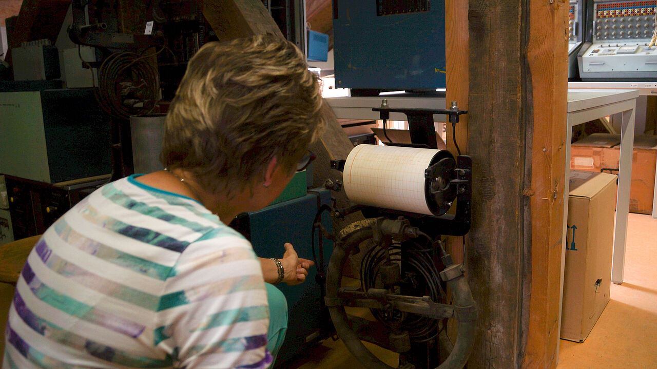 Eine Frau hockt mit dem Rücken zum Betrachter vor einer alten Maschine und erklärt, wie sie funktioniert